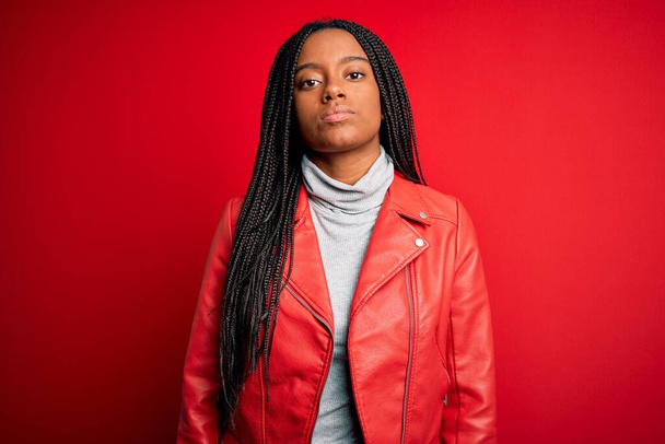 Jeune femme afro-américaine vêtue d'une veste en cuir cool sur fond rouge isolé Détendu avec une expression sérieuse sur le visage. Un regard simple et naturel sur la caméra
. - Photo, image