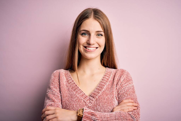 Jonge mooie roodharige vrouw draagt casual trui over geïsoleerde roze achtergrond vrolijk gezicht glimlachend met gekruiste armen kijkend naar de camera. Positief persoon. - Foto, afbeelding