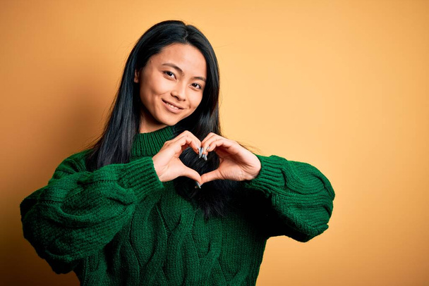 Young beautiful chinese woman wearing green sweater over isolated yellow background smiling in love showing heart symbol and shape with hands. Romantic concept. - Fotó, kép