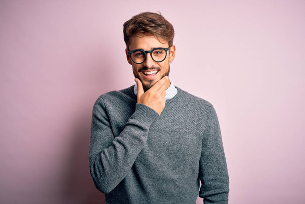 Mladý pohledný muž s vousy a brýlemi a svetrem, stojící nad růžovým pozadím, se sebevědomě usmívá na kameru se zkříženýma rukama a rukou zvednutou na bradě. Pozitivní myšlení. - Fotografie, Obrázek