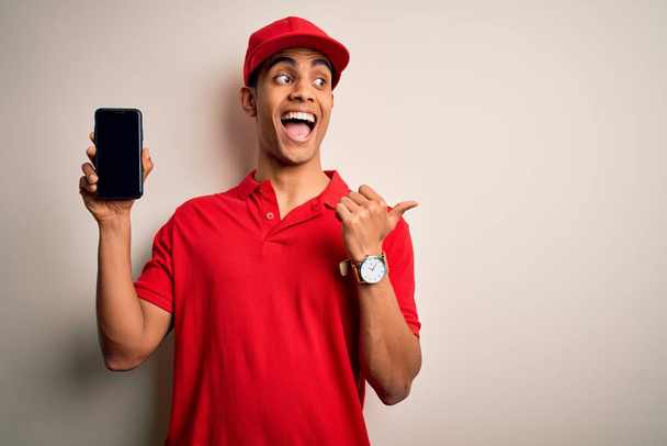 Молодий красивий афроамериканський чоловік доставки тримає смартфон, показуючи екранний додаток, вказуючи і показуючи з великим пальцем в сторону з щасливим обличчям посміхаючись
 - Фото, зображення