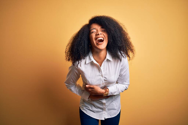 Jeune belle femme afro-américaine élégante avec des cheveux afro debout sur fond jaune souriant et riant fort à haute voix parce que drôle blague folle avec les mains sur le corps
. - Photo, image