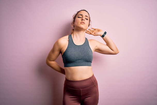 Young beautiful blonde sportswoman doing sport wearing sportswear over pink background Suffering of neck ache injury, touching neck with hand, muscular pain - Foto, Imagem