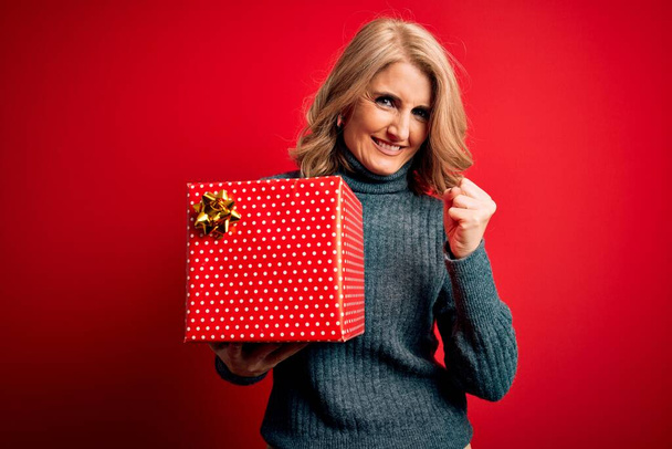 Middle age beautiful blonde woman holding birthday gift over isolated red background screaming proud and celebrating victory and success very excited, cheering emotion - Foto, Imagem