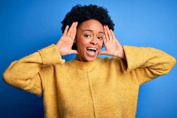 Young beautiful African American afro woman with curly hair wearing yellow casual sweater Smiling cheerful playing peek a boo with hands showing face. Surprised and exited - Photo, Image