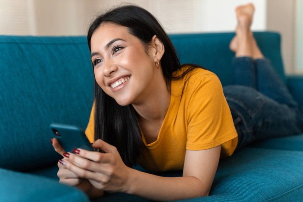 Photo of a beautiful happy pleased brunette young woman on sofa indoors at home using mobile phone. - Fotografie, Obrázek