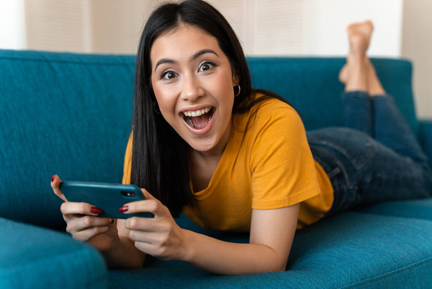 Photo of a beautiful surprised emotional pleased brunette young woman on sofa indoors at home using mobile phone. - Фото, зображення