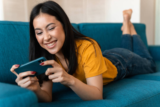 Photo of a beautiful cheery emotional pleased brunette young woman on sofa indoors at home play games by mobile phone. - Foto, immagini
