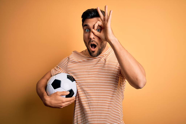 Beau joueur homme avec barbe jouant au football tenant ballon de football sur fond jaune faisant geste ok choqué par le visage surpris, oeil regardant à travers les doigts. Expression incrédule
. - Photo, image
