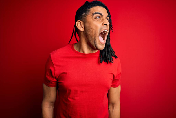 赤のカジュアルなTシャツを着たドレッドカジュアルなTシャツを着た恐ろしいロックを持つ若いハンサムなアフリカ系アメリカ人アフロの男は、怒りで叫んで、フラストレーションと激怒叫んでいます。激怒と積極的な概念. - 写真・画像