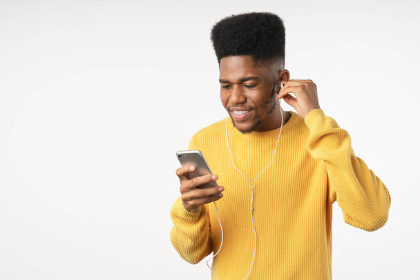 Portrait of smiling casual young african man with phone and earphones standing over white background - Zdjęcie, obraz