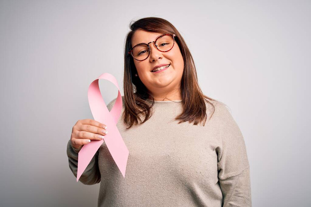 Beautiful plus size woman holding breast cancer awareness pink ribbon over isolated background with a happy face standing and smiling with a confident smile showing teeth - Photo, Image