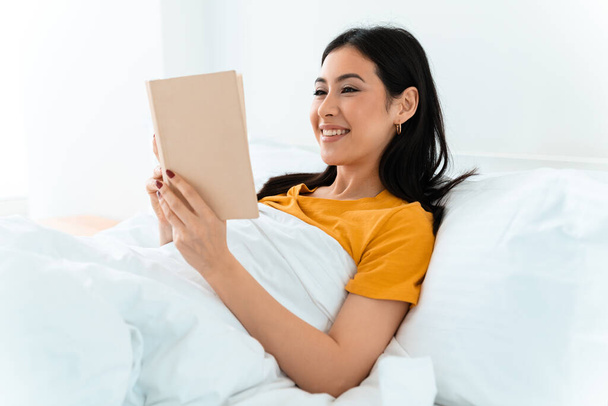 Image of a beautiful cheery positive young woman lies in bed indoors at home reading book. - Foto, Imagen