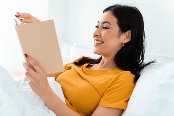 Εικόνα από μια όμορφη χαρούμενη θετική νεαρή γυναίκα βρίσκεται στο κρεβάτι σε εσωτερικούς χώρους στο σπίτι διαβάζοντας το βιβλίο. - Φωτογραφία, εικόνα