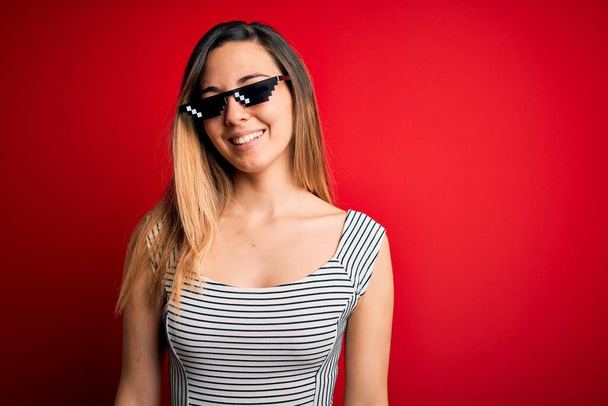 Junge schöne brünette Frau mit lustigen Schläger Leben Sonnenbrille über rotem Hintergrund mit einem glücklichen und kühlen Lächeln im Gesicht. Glücklicher Mensch. - Foto, Bild