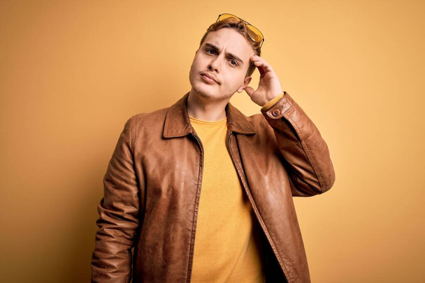 Young handsome redhead man wearing casual leather jacket over isolated yellow background confuse and wondering about question. Uncertain with doubt, thinking with hand on head. Pensive concept. - Photo, image