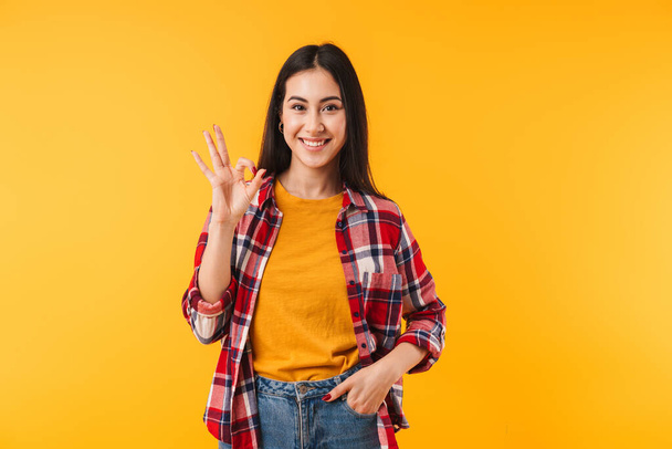 Image of joyful attractive woman in plaid shirt smiling and showing ok sign isolated over yellow background - Fotoğraf, Görsel