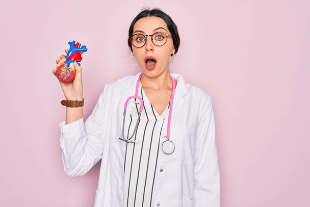 Young beautiful cardiologist woman with blue eyes wearing stethoscope holding heart scared in shock with a surprise face, afraid and excited with fear expression - Fotografie, Obrázek