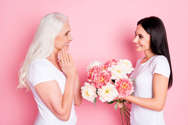 For you mommy. Profile photo of cheerful aged mother young daughter ladies spend time together giving fresh flowers bunch wear white t-shirts jeans isolated pastel pink color background - Foto, afbeelding