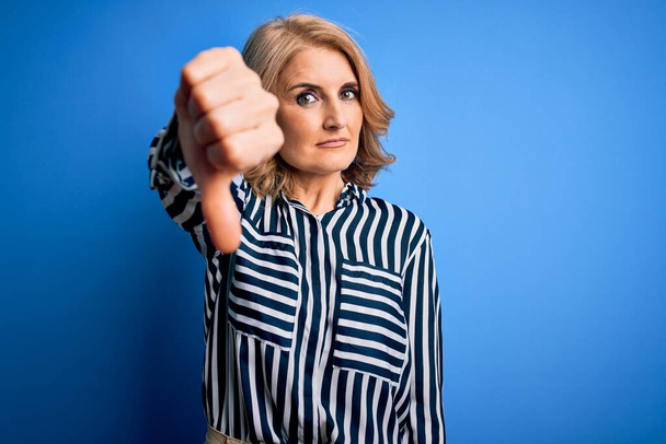 Middle age beautiful blonde woman wearing casual striped shirt standing over blue background looking unhappy and angry showing rejection and negative with thumbs down gesture. Bad expression. - Foto, Bild