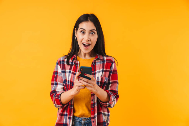 Image of surprised attractive woman in plaid shirt typing on cellphone isolated over yellow background - Foto, afbeelding