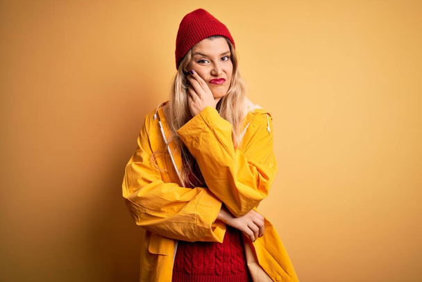 Young beautiful blonde woman wearing raincoat and wool cap over isolated yellow background thinking looking tired and bored with depression problems with crossed arms. - Photo, Image
