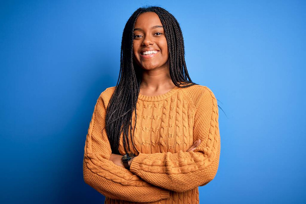 Jonge Afro-Amerikaanse vrouw die een casual gele trui draagt die over een blauwe geïsoleerde achtergrond staat te glimlachen met gekruiste armen die naar de camera kijken. Positief persoon. - Foto, afbeelding