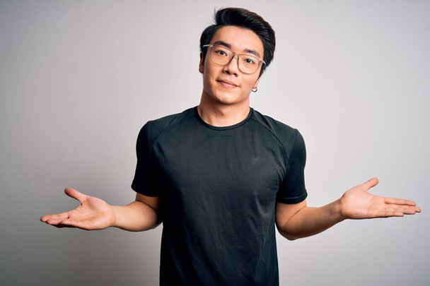 Jeune bel homme chinois portant un t-shirt noir et des lunettes sur fond blanc expression vide et confus avec les bras et les mains levées. Concept de doute
. - Photo, image