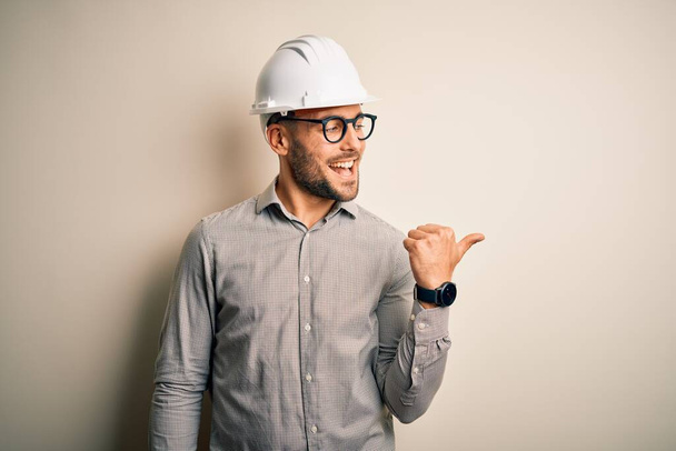 Молодой архитектор в защитном шлеме строителя на изолированном фоне улыбается со счастливым лицом и указывает на сторону с большим пальцем вверх
. - Фото, изображение