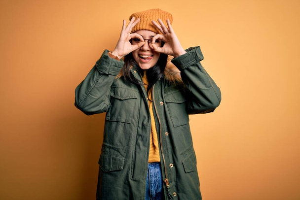 Young brunette woman wearing glasses and winter coat with hat over yellow isolated background doing ok gesture like binoculars sticking tongue out, eyes looking through fingers. Crazy expression. - Foto, immagini