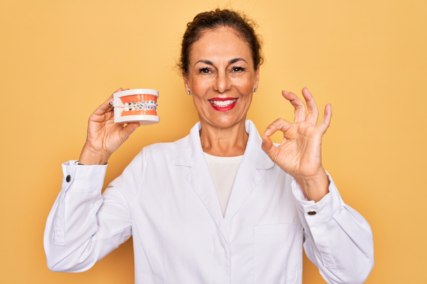 Старша жінка-стоматолог середнього віку, яка тримає зубний протез ортодонтії з брекетами інопланетянин робить знак з пальцями, відмінний символ
 - Фото, зображення