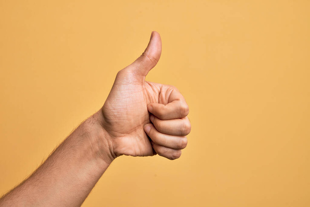 Hand of caucasian young man showing fingers over isolated yellow background doing successful approval gesture with thumbs up, validation and positive symbol - Photo, Image