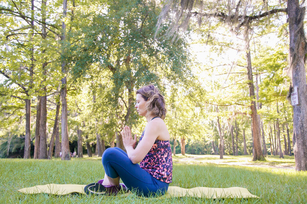 Ragazza seduta sull'erba nel parco e che si tiene per mano in una posa di preghiera al sole al tramonto. Il concetto di uno stile di vita sano, yoga e meditazione
. - Foto, immagini