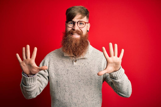 Bonito irlandês ruiva homem com barba vestindo camisola casual e óculos sobre fundo vermelho mostrando e apontando para cima com os dedos número dez enquanto sorri confiante e feliz
. - Foto, Imagem