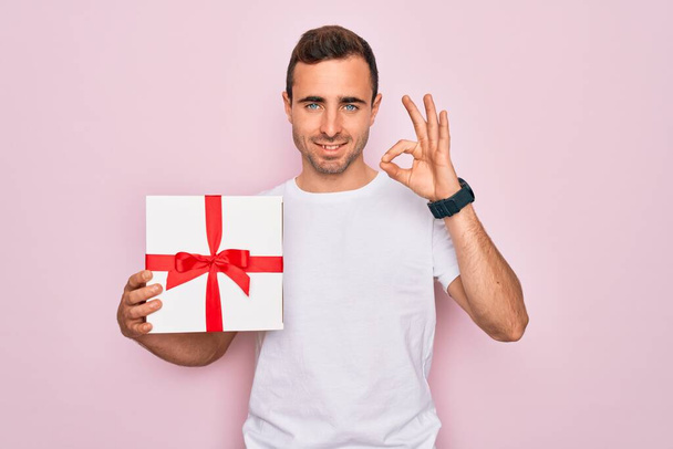 Jeune bel homme aux yeux bleus tenant cadeau d'anniversaire sur fond rose isolé faisant signe ok avec les doigts, excellent symbole
 - Photo, image