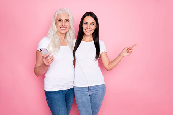 Photo of pretty old mother young daughter two ladies hold telephone hugging direct finger side empty space wear casual white t-shirts jeans isolated pastel pink color background - 写真・画像