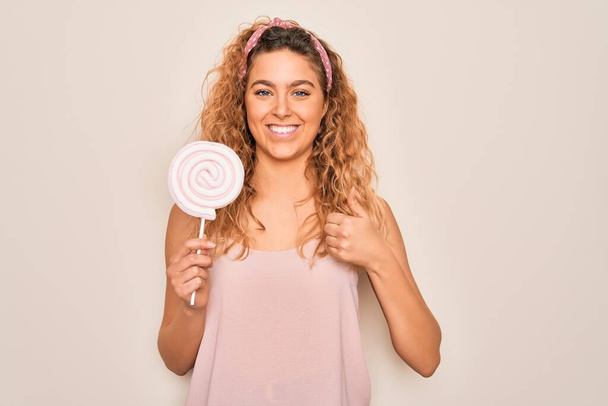 Young beautiful blonde woman with blue eyes eating sweet candy lollipop over pink background happy with big smile doing ok sign, thumb up with fingers, excellent sign - Photo, Image