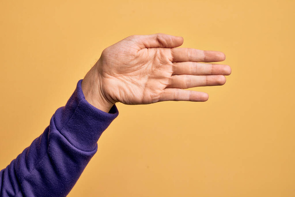 Hand of caucasian young man showing fingers over isolated yellow background stretching and reaching with open hand for handshake, showing palm - Fotoğraf, Görsel