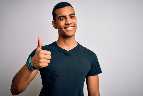 Junger gutaussehender afrikanisch-amerikanischer Mann in lässigem T-Shirt, der vor weißem Hintergrund steht und mit der Hand fröhlich Daumen nach oben gestikuliert. Zustimmender Blick in die Kamera zeigt Erfolg. - Foto, Bild