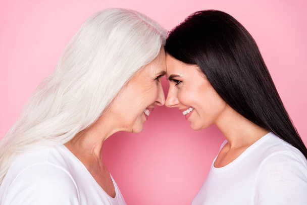 Closeup profile photo of pretty old mother young daughter ladies stand opposite lean foreheads affectionate look eyes wear casual white t-shirts isolated pastel pink color background - Foto, imagen
