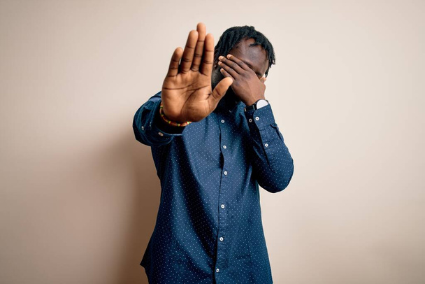 Mladý pohledný africký Američan v neformální košili, stojící nad bílým pozadím a zakrývající oči rukama a vykonávající gesto se smutným a ustrašeným výrazem. Ztrapněný a negativní koncept. - Fotografie, Obrázek