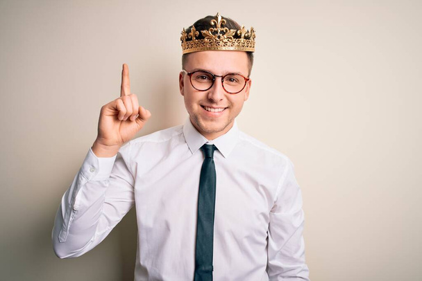 Young handsome caucasian business man wearing golden crown over isolated background showing and pointing up with finger number one while smiling confident and happy. - Fotografie, Obrázek
