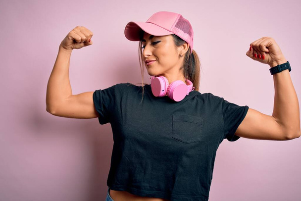 Молода красива фітнес спортивна жінка носить тренувальну шапку і навушники на рожевому фоні, показуючи м'язи рук, які посміхаються гордими. Концепція фітнесу
. - Фото, зображення