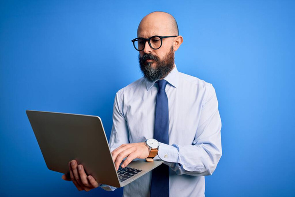Sakallı yakışıklı, kel bir iş adamı mavi arka planda dizüstü bilgisayar kullanıyor. - Fotoğraf, Görsel