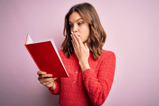 Молода блондинка студентка читає книгу над рожевим ізольованим фоновим обкладинкою ротом з рукою, шокованою соромом за помилку, вираження страху, налякана тишею, таємною концепцією
 - Фото, зображення