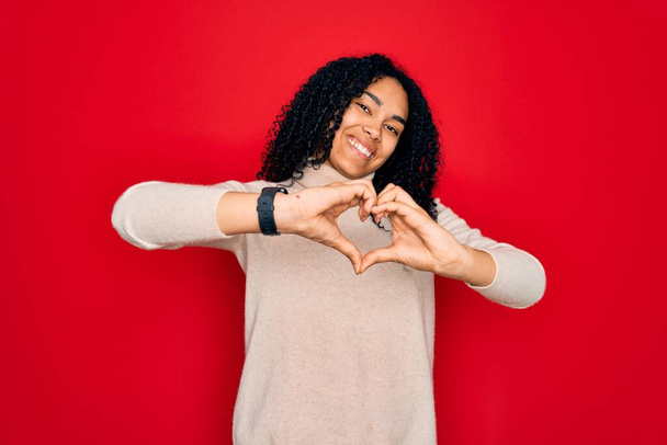 Mujer rizada afroamericana joven que usa suéter casual de cuello alto sobre fondo rojo sonriendo en amor haciendo forma de símbolo del corazón con las manos. Concepto romántico
. - Foto, Imagen
