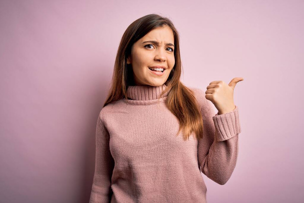 Beautiful young woman wearing turtleneck sweater over pink isolated background smiling with happy face looking and pointing to the side with thumb up. - Photo, Image
