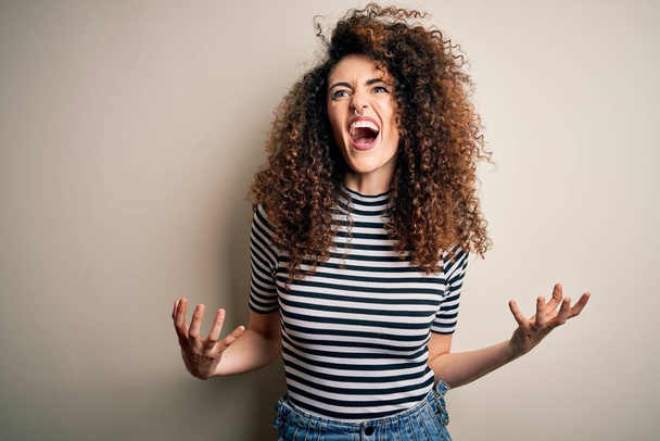 Young beautiful woman with curly hair and piercing wearing casual striped t-shirt crazy and mad shouting and yelling with aggressive expression and arms raised. Frustration concept. - Photo, image