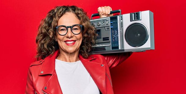 Middelbare leeftijd brunette hipster vrouw met retro muziek boombox over rood geïsoleerde achtergrond kijken positief en gelukkig staan en glimlachen met een zelfverzekerde glimlach tonen tanden - Foto, afbeelding