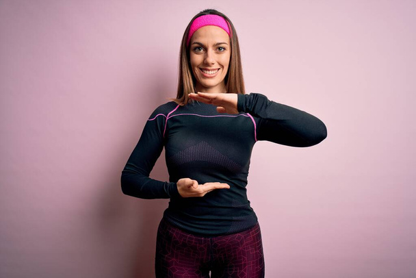 Jonge blonde fitnessvrouw dragen sport workout kleding over geïsoleerde achtergrond gebaren met handen tonen grote en grote maat teken, maatregel symbool. Lachend kijkend naar de camera. Meetconcept. - Foto, afbeelding